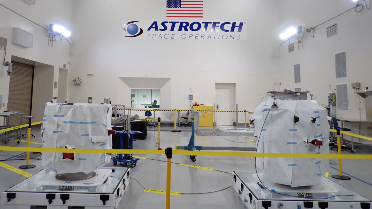 Two metal satellites wrapped in fabric sit on two carts behind yellow tape in a factory