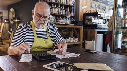 An older man who is an entrepreneur sits at a table in his shop and adds up receipts on a calculator.