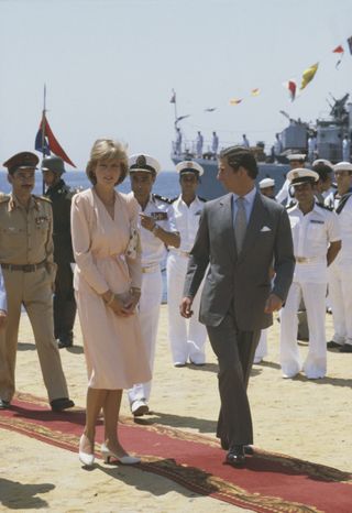 Princess Diana wearing a pink dress walking outside next to Prince Charles and a row of sailors in Egypt