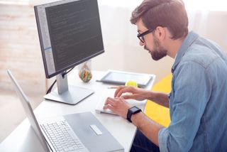 Person typing code on a keyboard with a large monitor displaying code, a laptop, and a tablet on the desk.