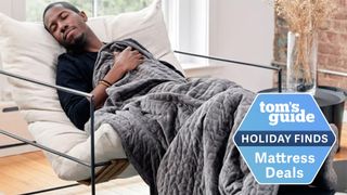 Image shows a man napping under a grey Gravity Weighted Blanket during the daytime, with a blue Holiday Mattress Deals badge overlaid on the image