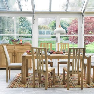 six seater wooden dining table and chairs on top of a patterned rug, with fruit bowls on the table, and a wooden sideboard up against the glass walls of a white conservatory
