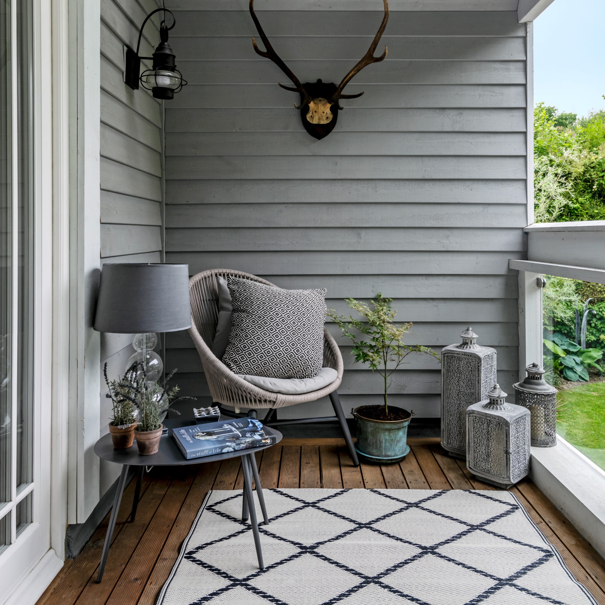 A balcony with panelled walls painted grey with a rattan chair in the corner, lantern lights on the floor and a table lamp on a coffee table