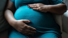 close up of a pregnant Black woman's belly. She wears a teal dress and has her hands gently placed on top and bottom of her stomach