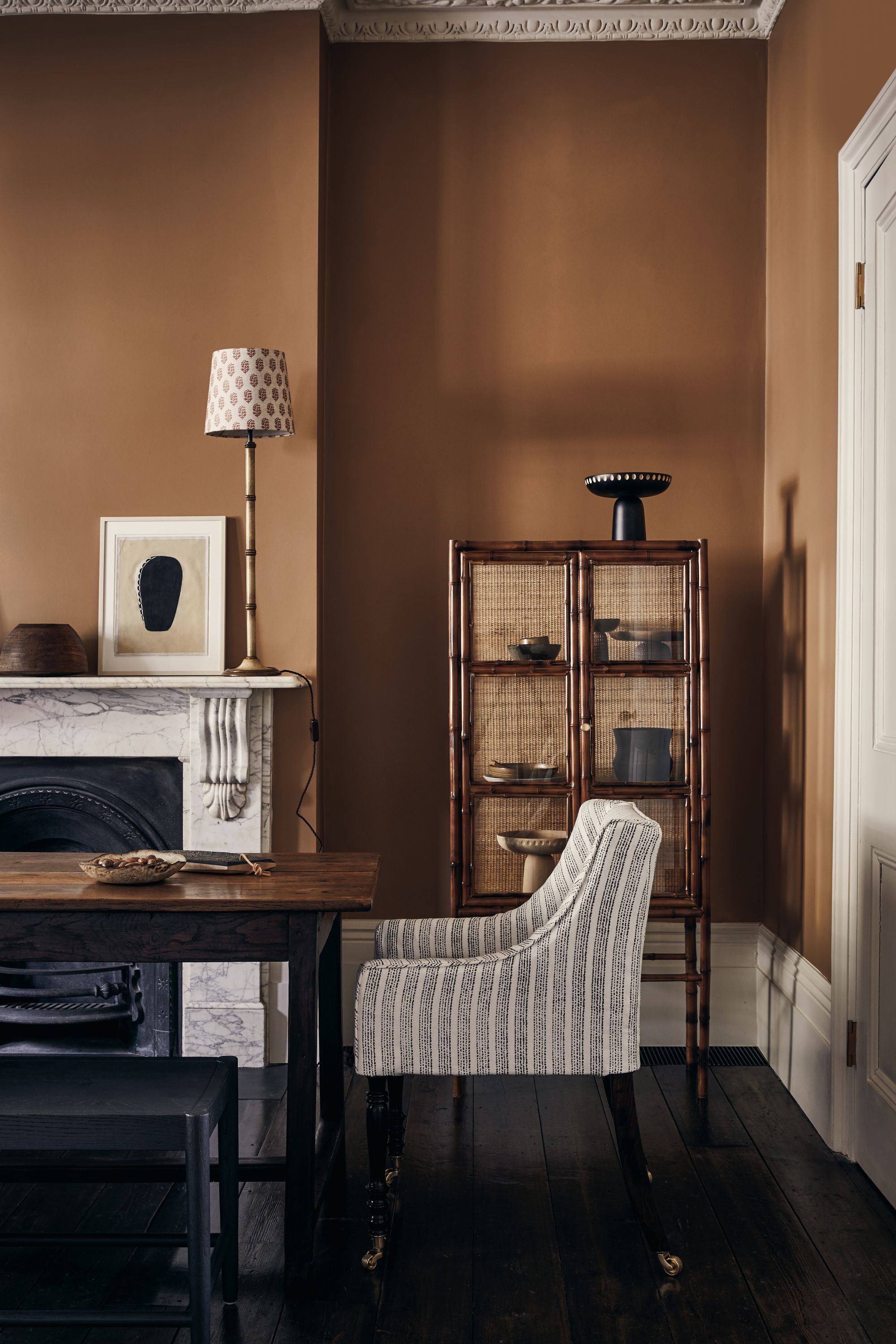 A dining room with brown walls, white cornicing, and a white and gray veined fireplace, a wooden dining table, black benches, and a gray and white striped armchair next to a display cabinet