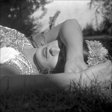 a woman (frida kahlo) wearing a flower crown lies in a grassy field while resting her arm over her eyes