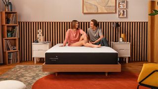 The best Cocoon by Sealy mattress sales, deals and discount codes: a couple sitting on the Cocoon by Sealy Chill Hybrid Mattress in a bedroom on a wooden frame 