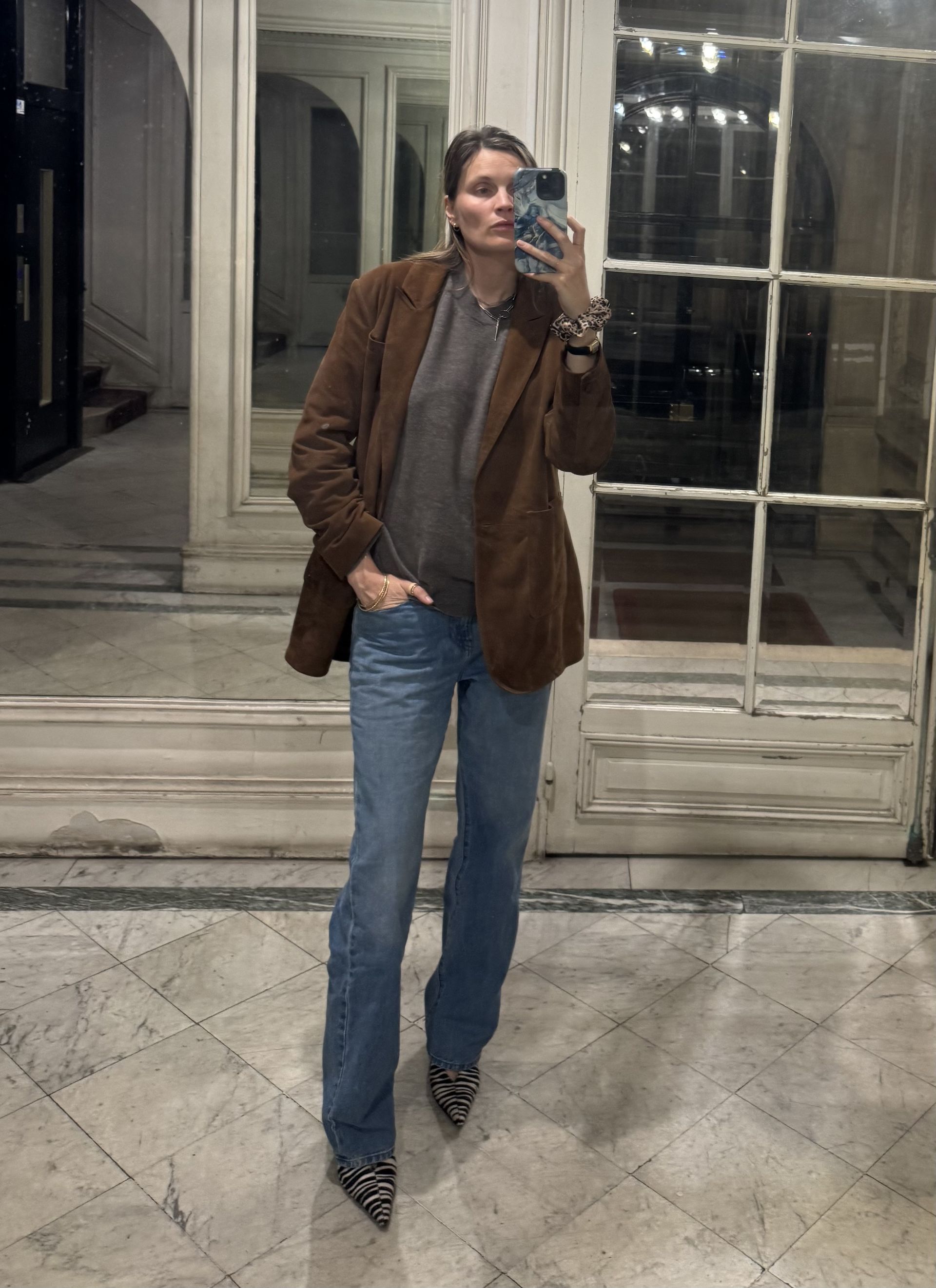French writer and editor Eugenie Trochu poses for a mirror selfie wearing a brown suede leather blazer jacket, gray top, bootcut jeans, and zebra-print heels