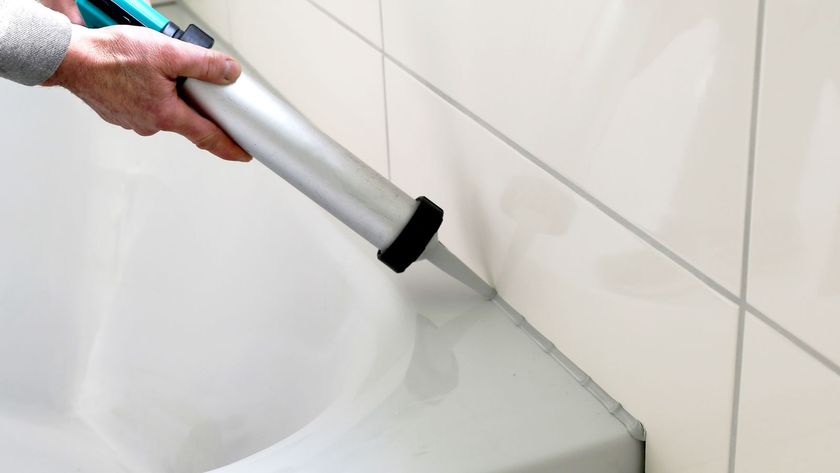Person holding a silver caulk gun adding grey sealant between white bath and cream tiles