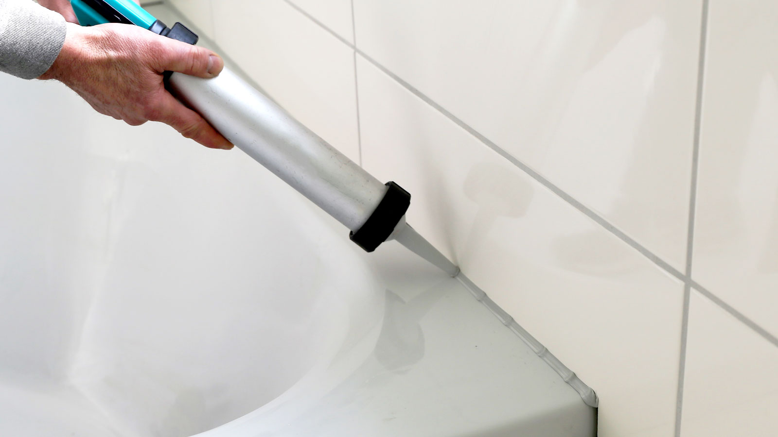 Person holding a silver caulk gun adding grey sealant between white bath and cream tiles