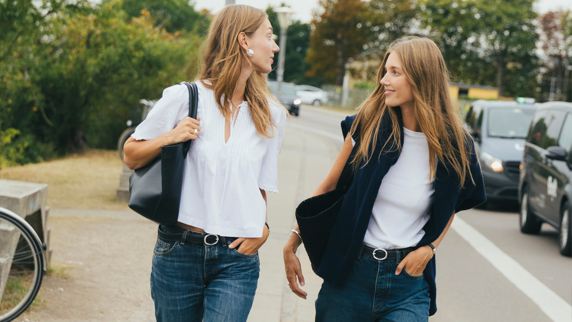Two woman walk and pose. One wears a short sleeve white blouse with jeans and the other wears a white tee, blazer, and jeans. 