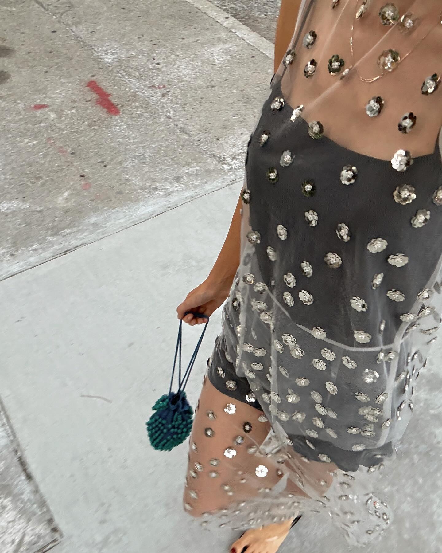 NYC based style influencer Courtney Grow takes an outfit selfie wearing a sheer rhinestone covered dresses over a black mini slip dress with a green beaded Bode bag, and clear jelly flip-flop sandals