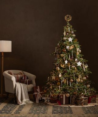 A Christmas tree beside a gray armchair against a dark brown wall.