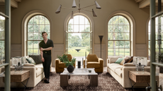 a large living room with three large windows, two couches, two accent chairs, a large coffee table, and a man standing to the left side of the room