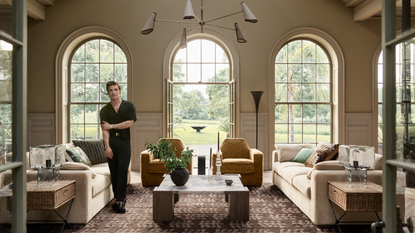 a large living room with three large windows, two couches, two accent chairs, a large coffee table, and a man standing to the left side of the room