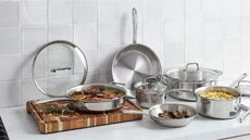 All-Clad Stainless Steel D3 cookware on a shite kitchen counter, with a butcher block cutting board. 
