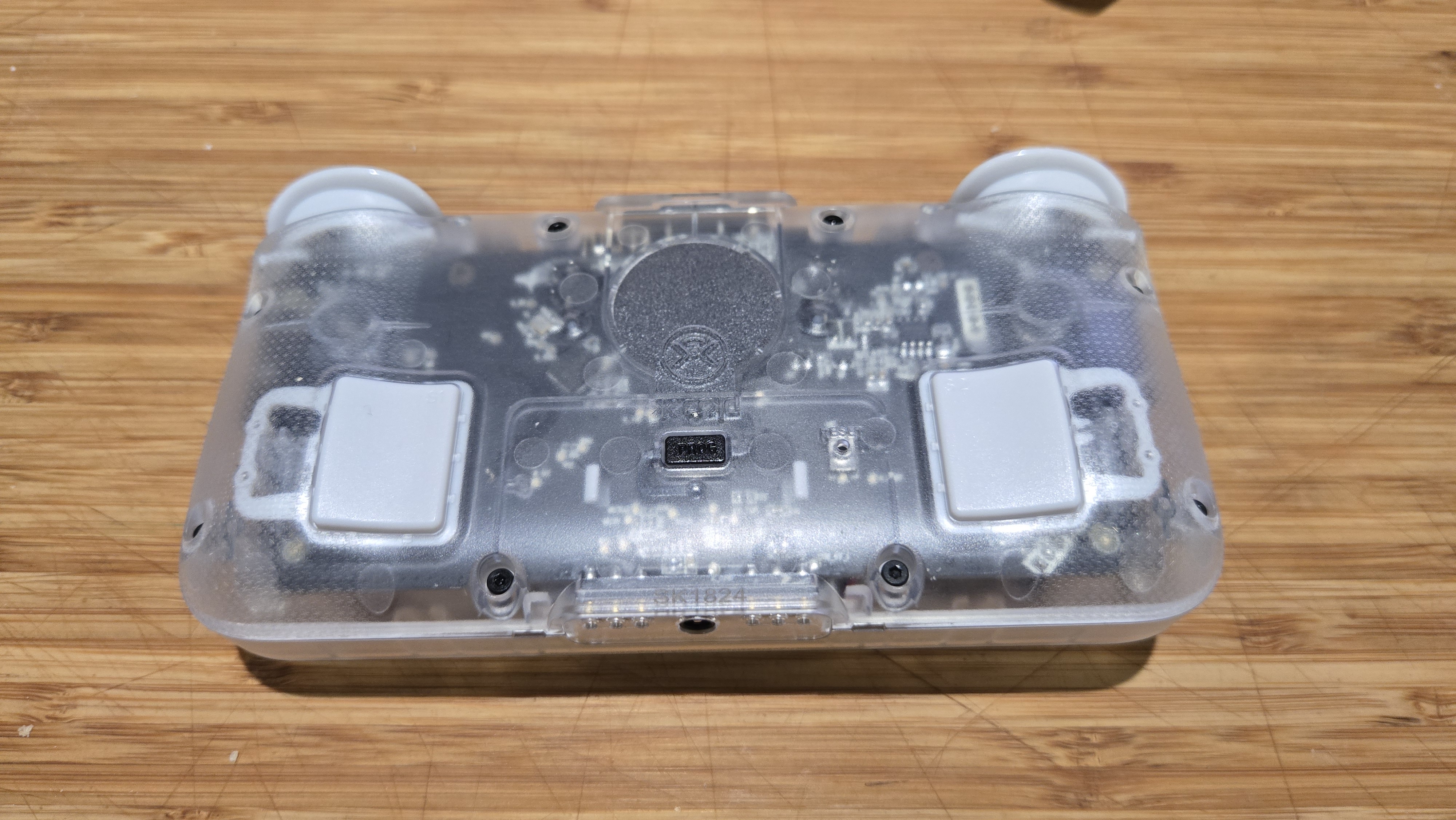 A Classic Clear edition of the CRKD NEO S rectangular controller. It is placed upon a wooden countertop in such a manner that the underside of the controller is facing up, towards the viewer. The body of the controller is made out of a cloudy clear plastic that allows the viewer to see the internals.