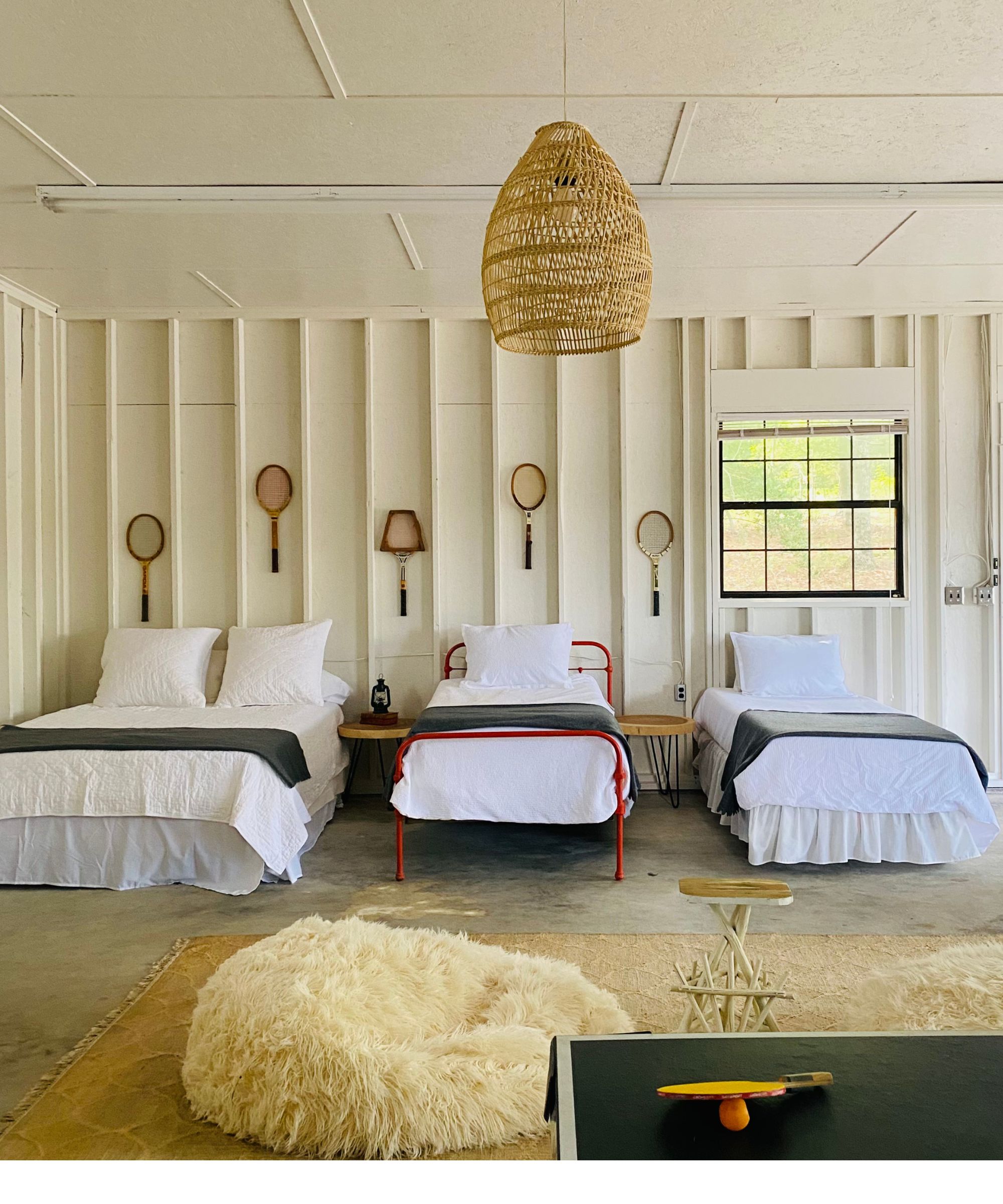 Sleeping porch with white panelling, three single beds and vintage tennis rackets on wall with fluffy rug and rattan light