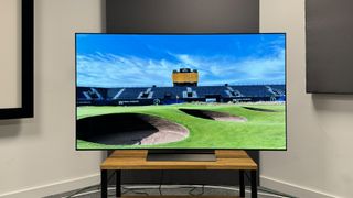 65-inch LG C4 TV photographed straight-on on a wooden stand. On the screen is an image of a golf course.