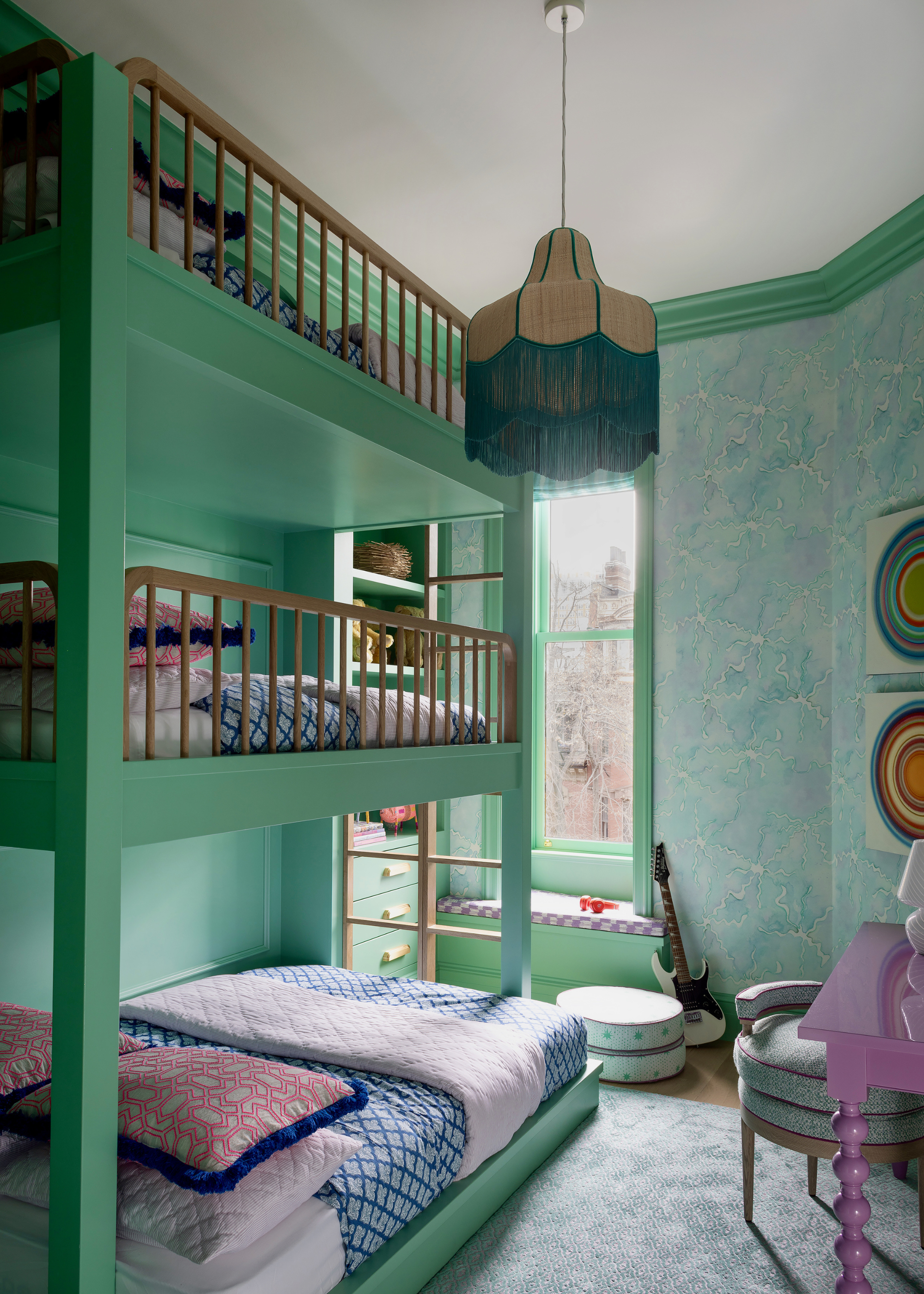 A modern children's bedroom with a three-tiered bunk bed painted in a vibrant green which is brought across the trims and window frame in the room; the back wall is covered in a light teal and green patterned wallpaper and a vibrant purple bobbin table is just out of view