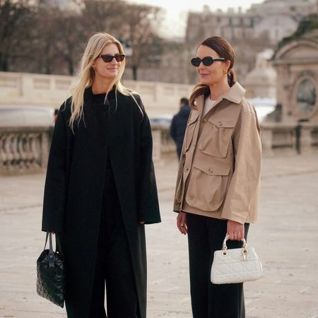 Two women pose while speaking to each other in Paris. The one on the left is wearing an all black outfit with sunglasses and an Issey Miyake handbag. The woman on the left is wearing a khaki jacket and black pants with a white handbag and sunglasses. 