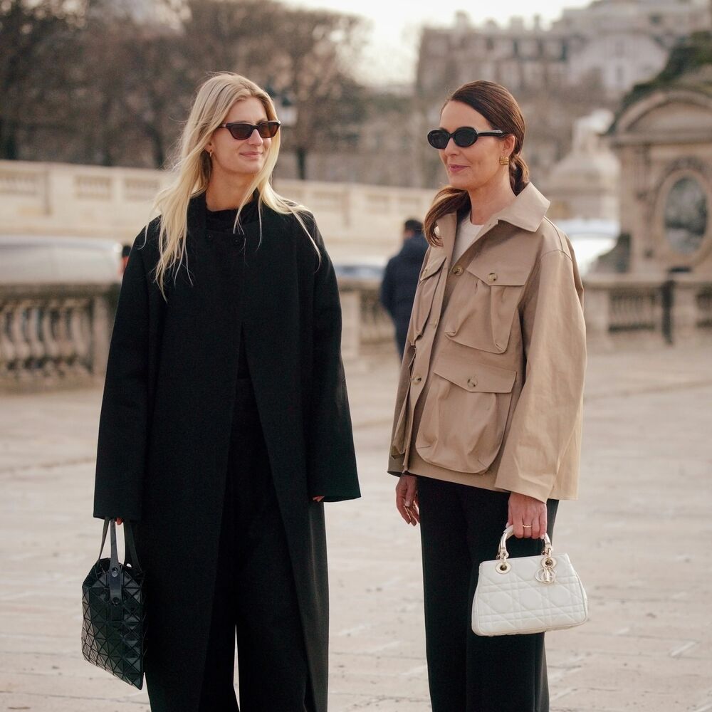 Two women pose while speaking to each other in Paris. The one on the left is wearing an all black outfit with sunglasses and an Issey Miyake handbag. The woman on the left is wearing a khaki jacket and black pants with a white handbag and sunglasses. 