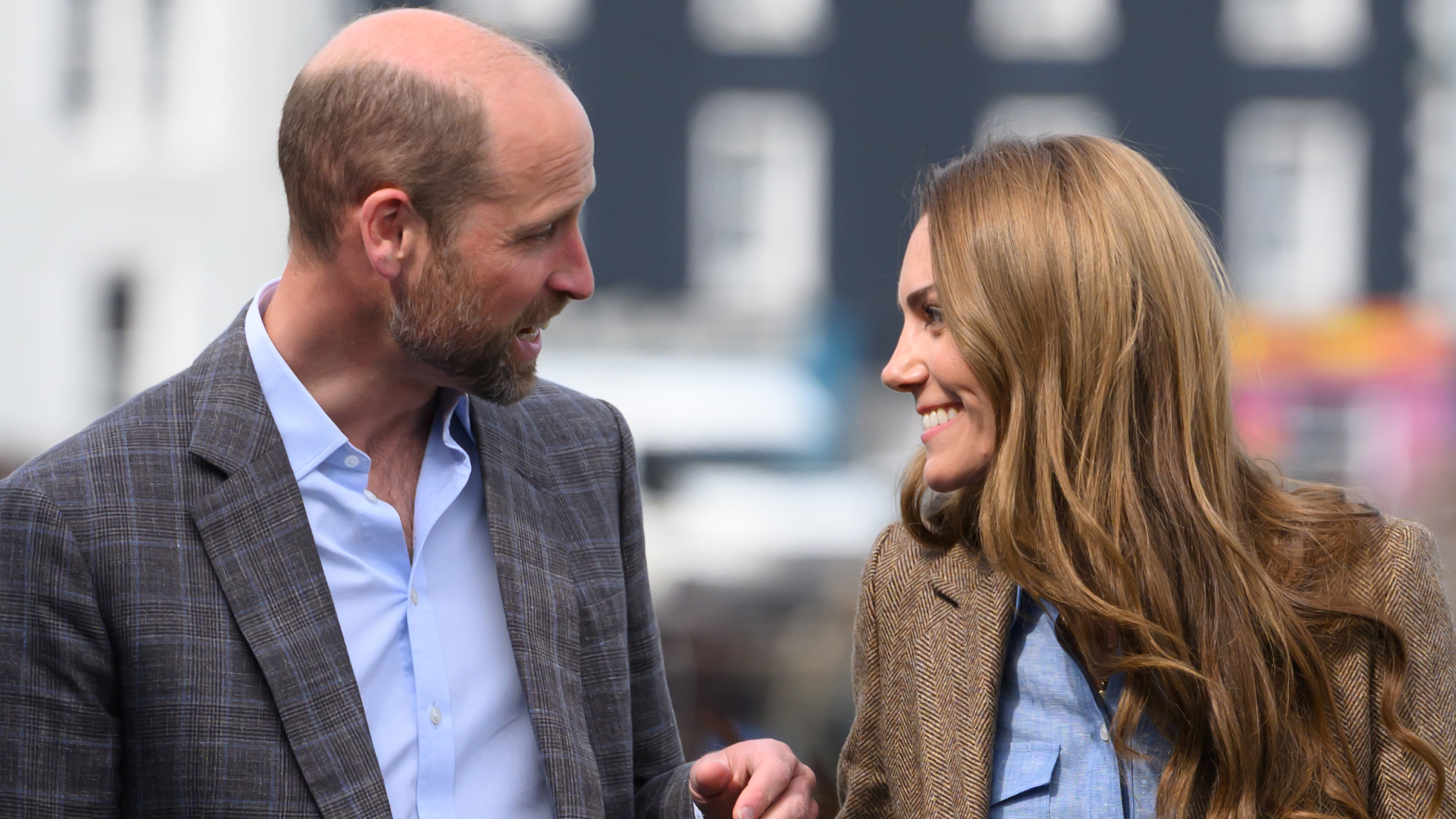 Prince William and Kate Middleton wearing blazers and blue shirts looking at each other and smiling