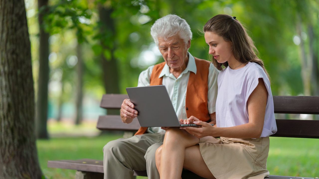 A granddaughter helps her grandfather with his laptop while sitting on a park bench.