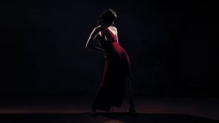 A woman in a red dress stands gracefully in a dimly lit environment, creating a striking contrast with the darkness