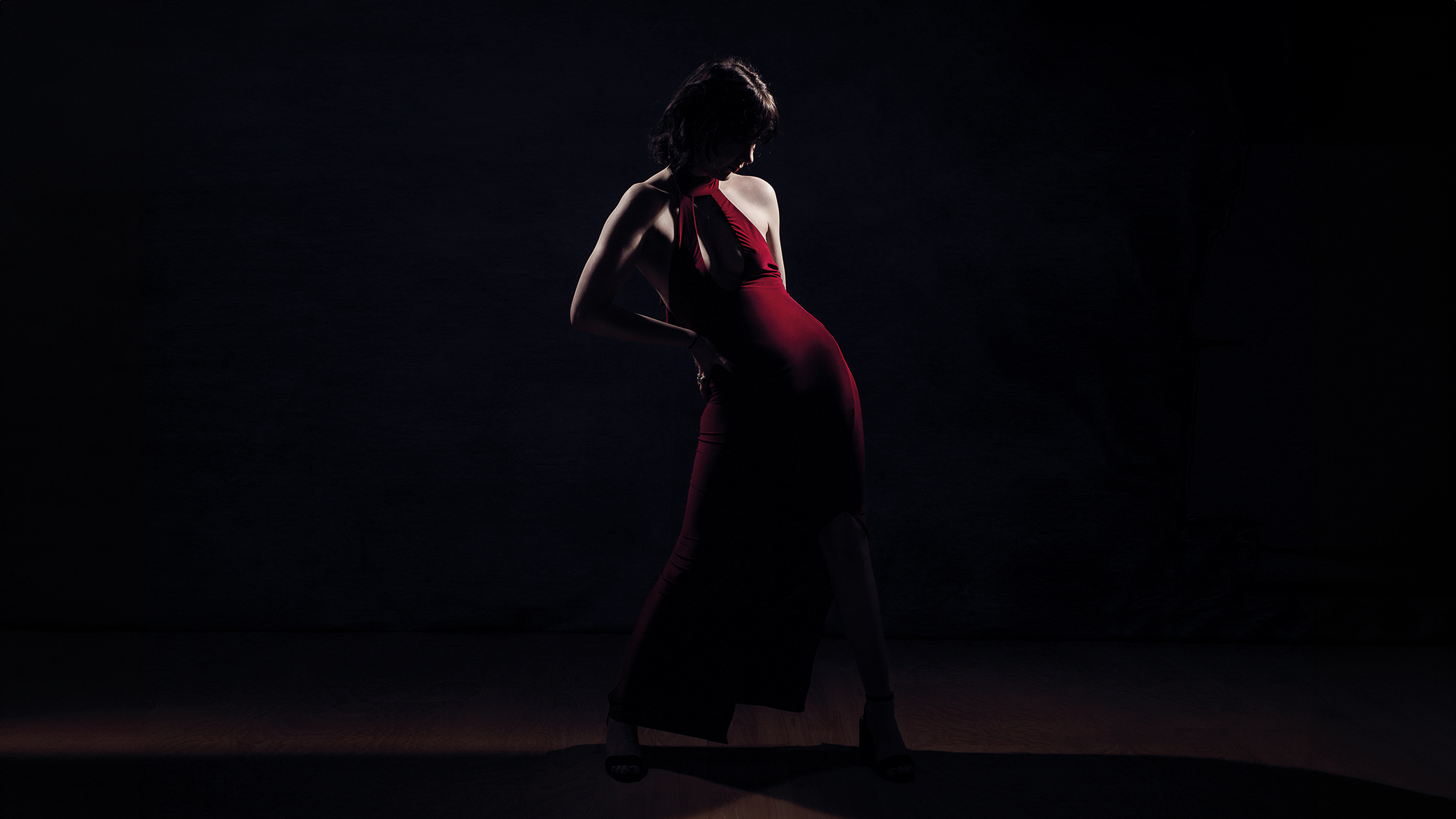 A woman in a red dress stands gracefully in a dimly lit environment, creating a striking contrast with the darkness