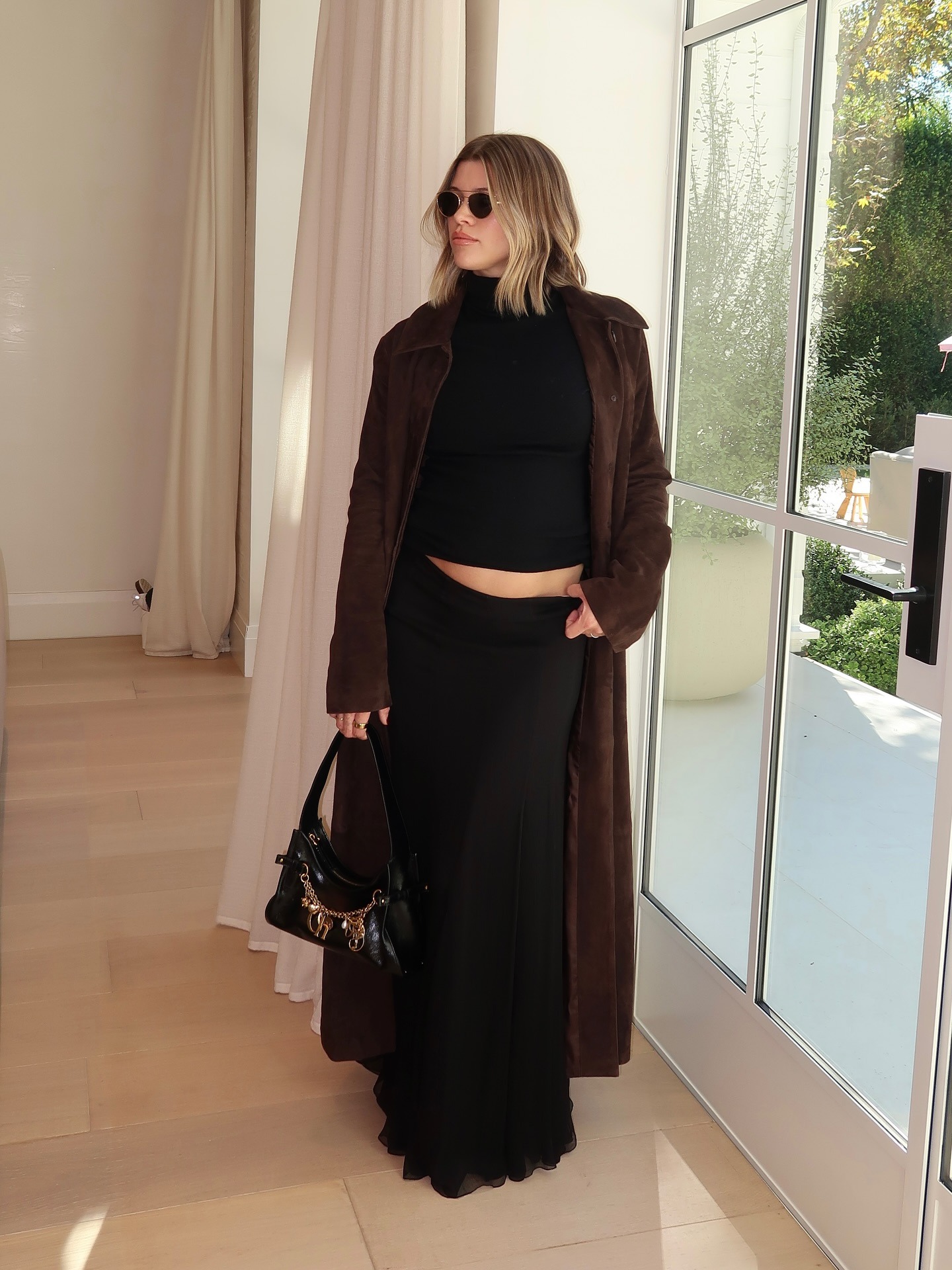 Sofia Richie Grainge poses indoors wearing a black turtleneck top with a black maxi skirt, a brown suede trench coat and a black bag with chain detailing.