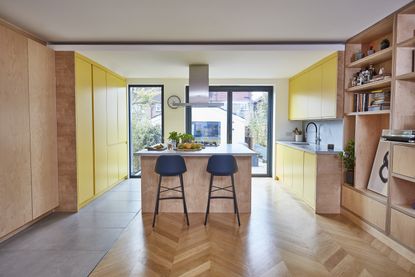 Plywood kitchen with yellow units and an island with dark blue and black bar stools