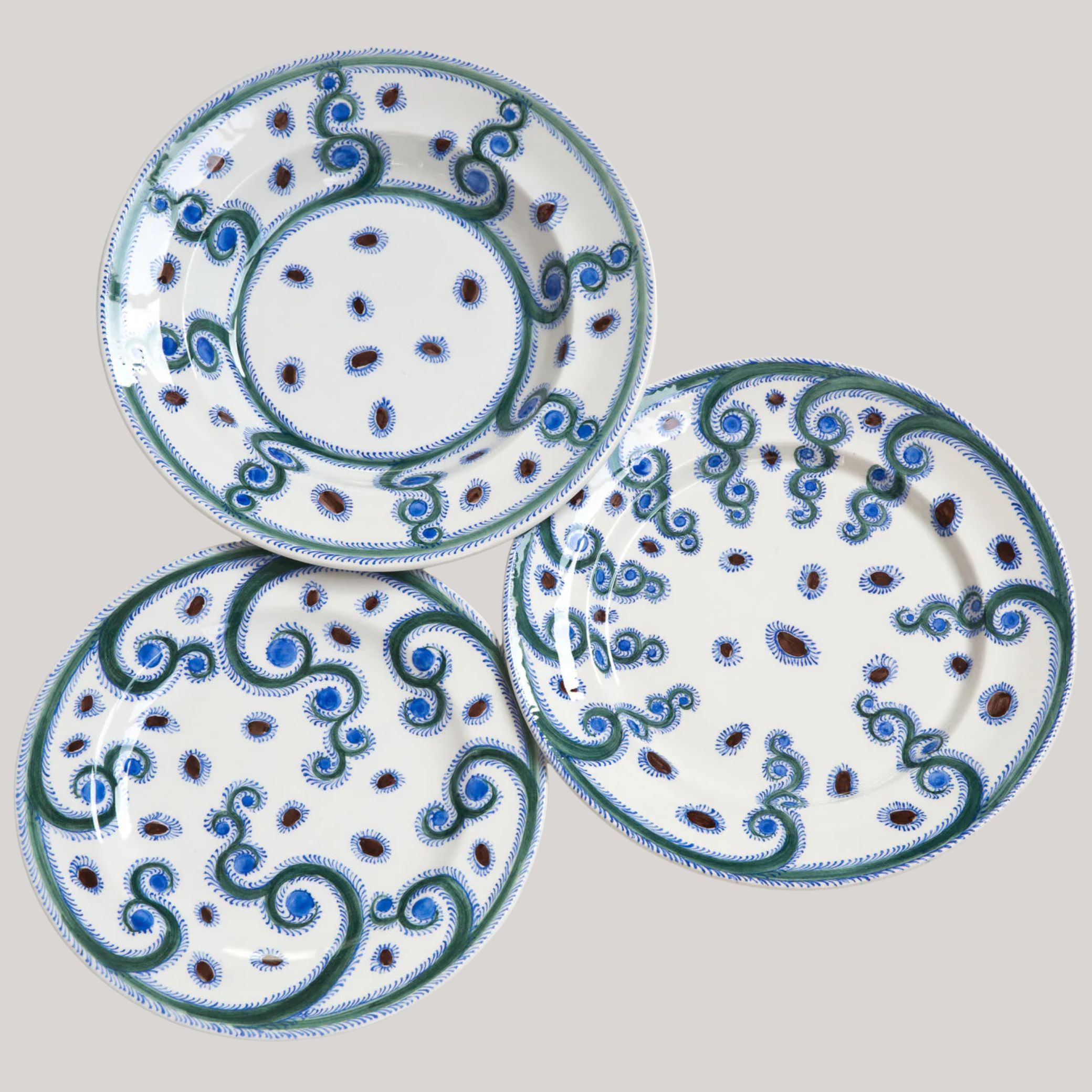 Three white porcelain plates painted with abstract, striking motifs in the tones of blue, ocean green, and dark red sit against a pale gray background.
