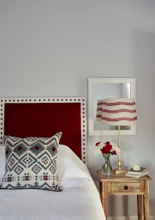 Edge of bed with headboard, bedside table with lamp and mirror behind, and white wall