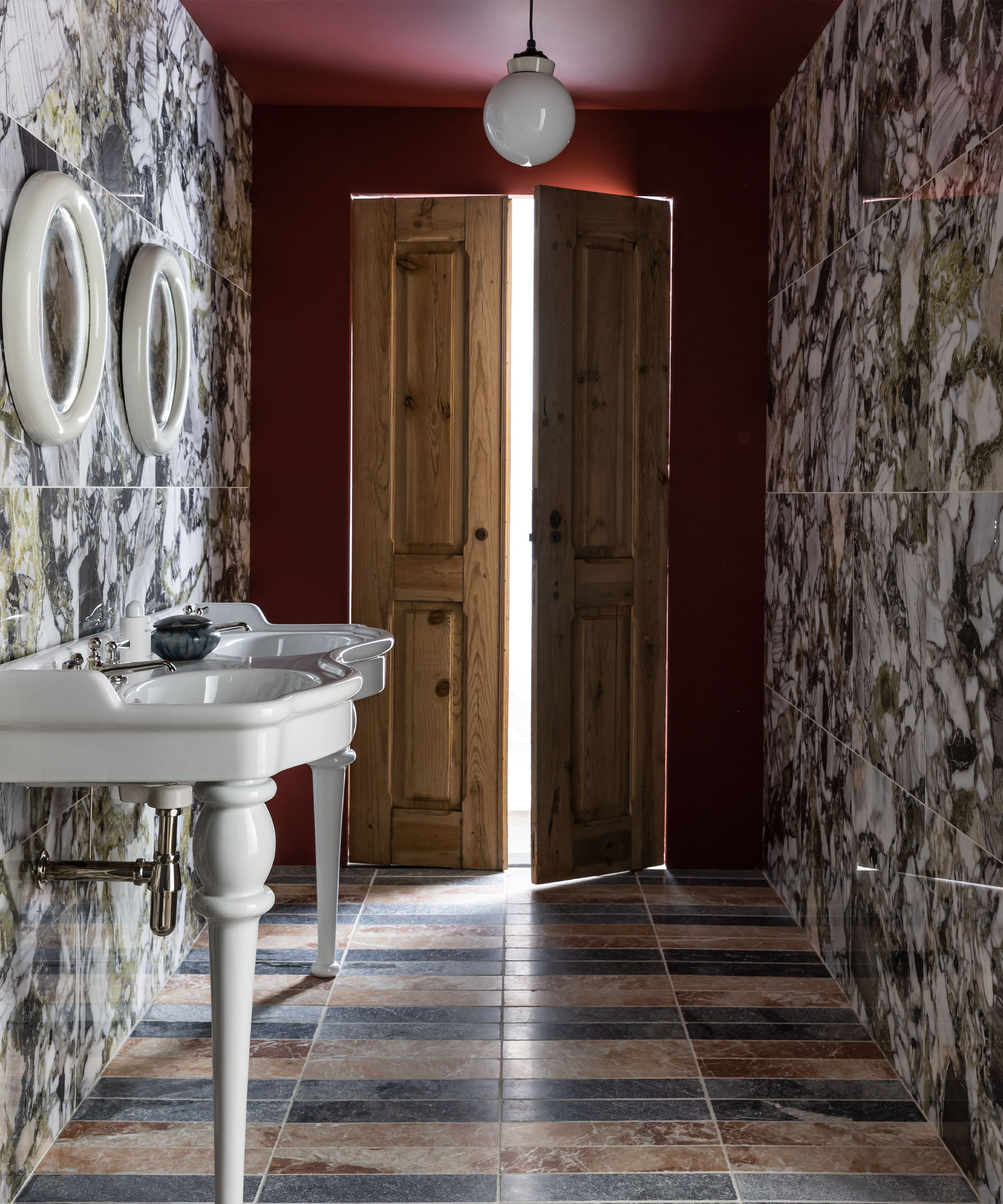 Bathroom with tiled flooring in black and brown floor tiles, multi-colored marble wall tiles, dark red painted wall and ceiling, traditional wooden shutter doors, twin basin sink with two rounded white mirrors mounted above, rounded opal glass pendant light