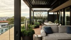 A balcony with a glass railing, wood panelled flooring, potted foliage and comfy seating