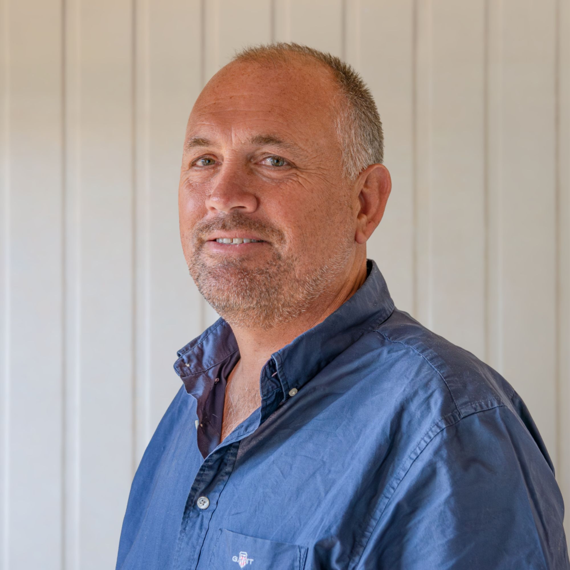 Man with grey stubble and cropped hair, wearing navy shirt, standing side-on against a panelled cream wall.