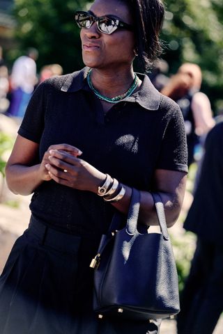 Nikki Ogunnaike wears a black knit polo shirt and trousers, a Hermés Picotin bucket bag, sunglasses, and a silver wrap-around Bulgari Serpenti watch at New York Fashion Week.