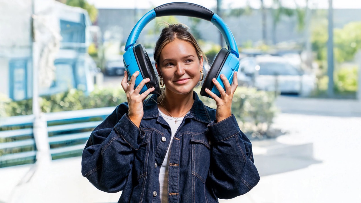 A woman holding a massively oversized pair of over-ear headphones &ndash; the earcups of which are the size of her head &ndash; as if about to put them on, with a wry smile on her face.