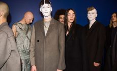Six male models wearing looks from the Rick Owens collection. One model is wearing a grey multi-layered piece with chain and straps. Another two models are wearing grey jackets and another three models are wearing dark coloured jackets. There are two models with painted white faces and a seventh person in the background with an afro. They are standing in front of a blue wall