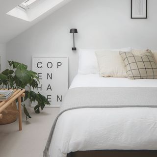 White loft bedroom with white bedding and cushions on it, with a white 'COPENHAGEN' print standing on the floor next to the bed