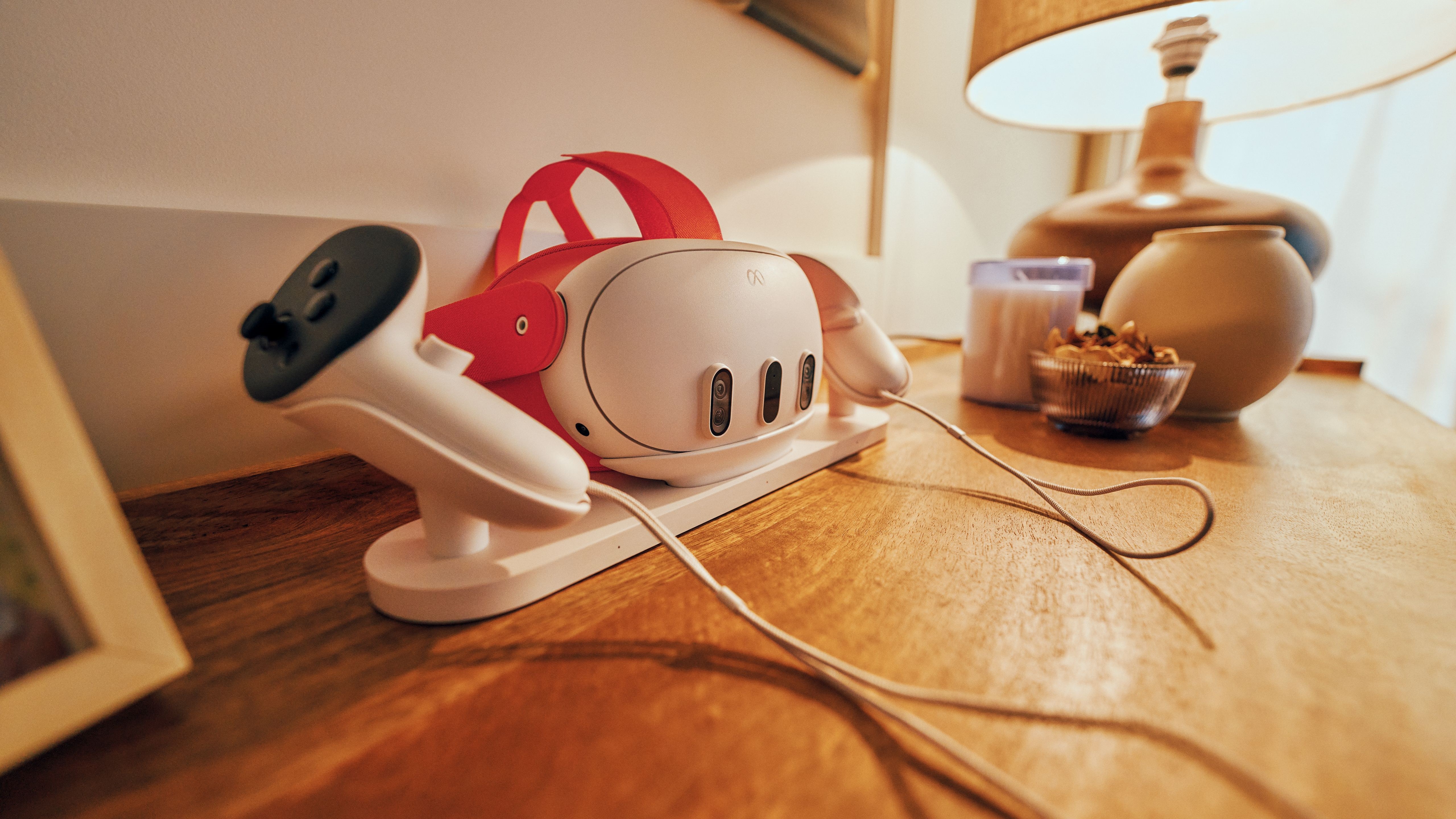 The Meta Quest 3 and its controllers in a charging station on a wooden top next to candles, a vase and a lamp