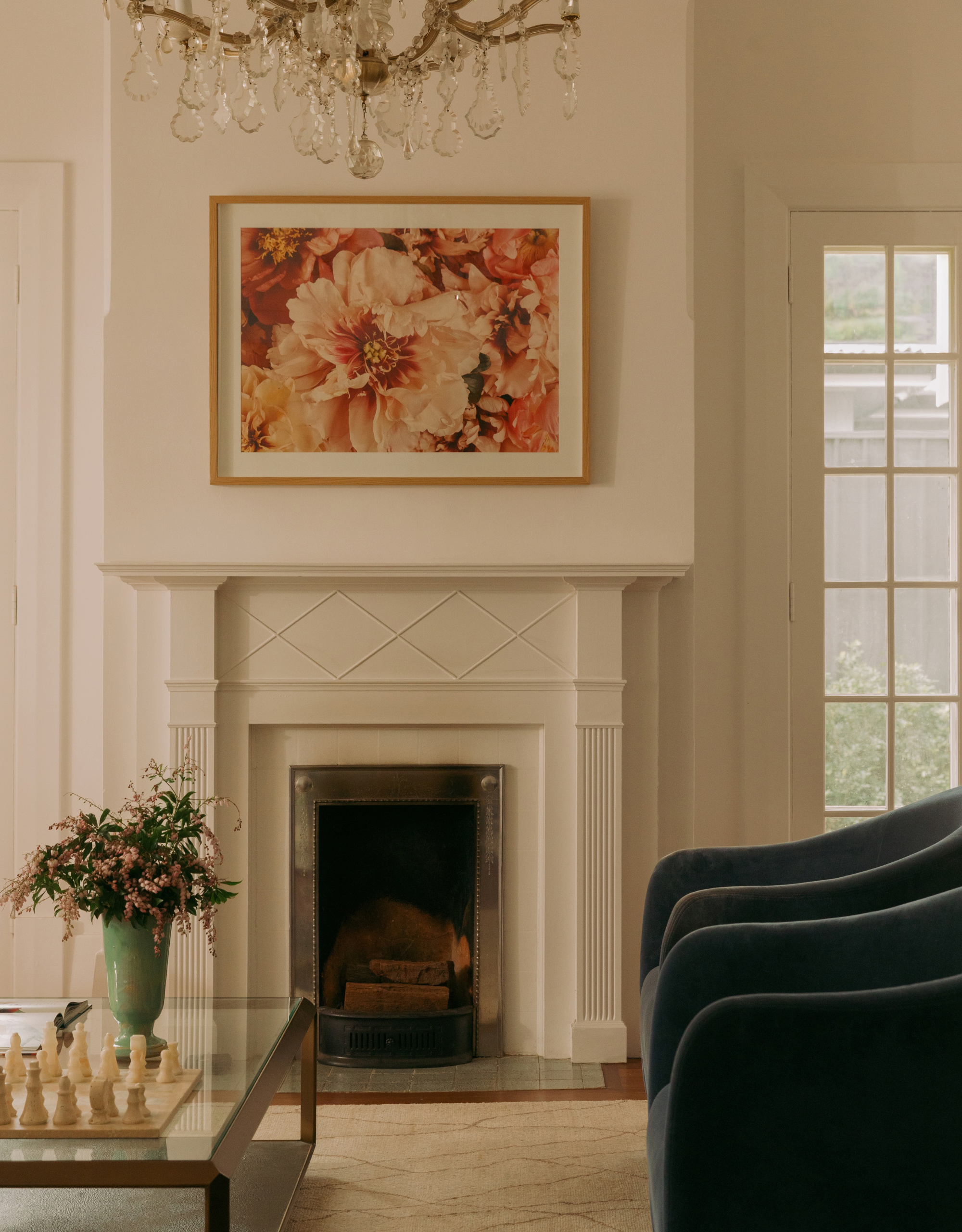 A white living room with a pair of dark blue armchairs, a glass coffee table, a vase of flowers, a chess set, a fireplace, a chandelier, and a framed floral print