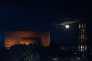 full moon rising over buildings in a partially cloudy sky.