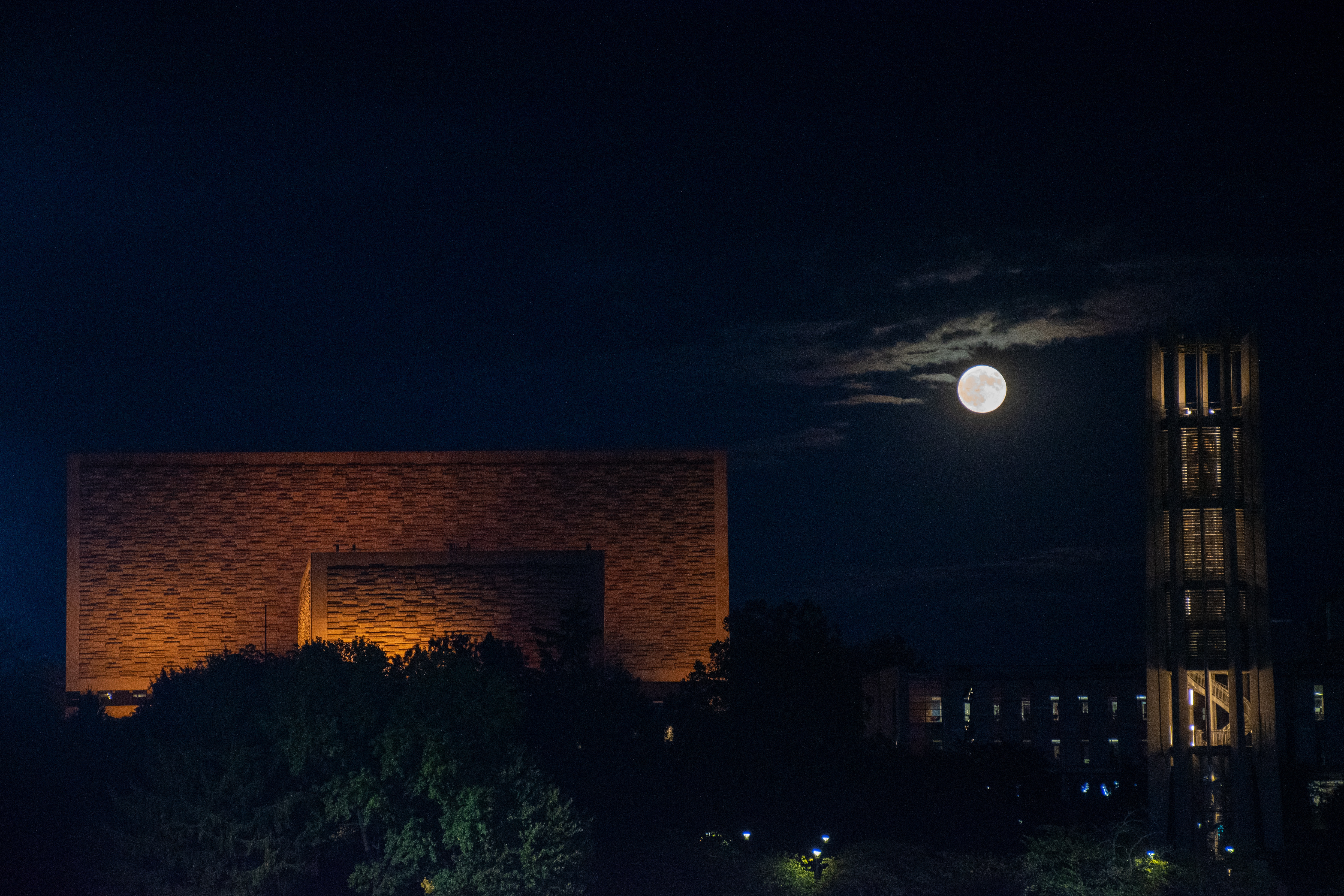 Full moon rises over buildings in a partly cloudy sky.