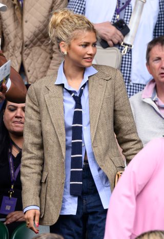 Zendaya in the stands at Wimbledon wearing a gray blazer with a blue button down shirt and a striped tie