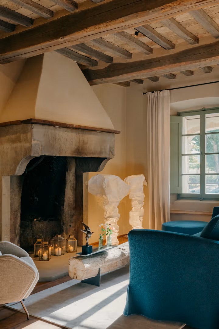 An heritage property with exposed wooden beams, a worn fireplace, and mint green-painted windows is decorated with sculptural design finds like crunched paper lamps, petrol green sofas, and rugs.