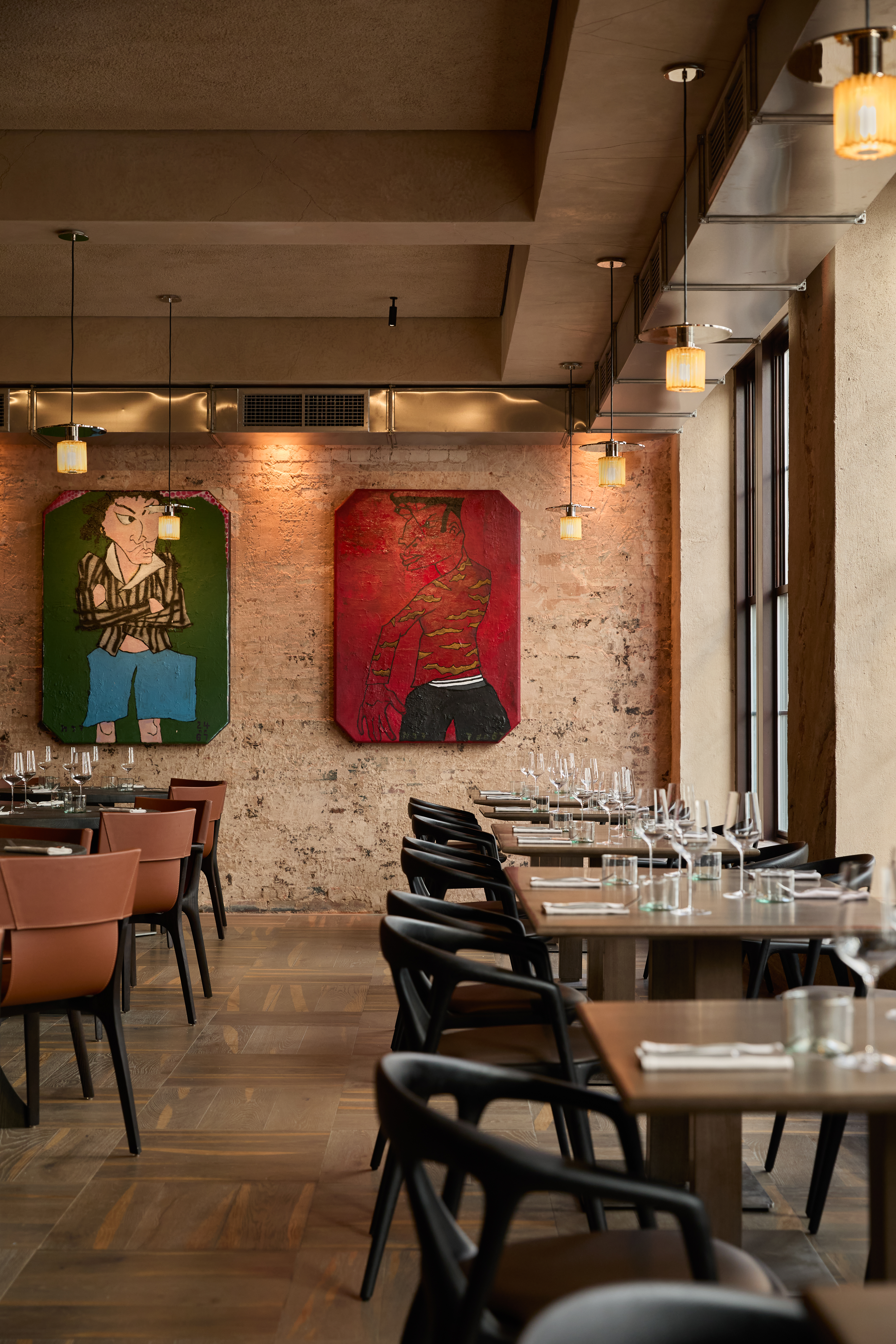 A minimalist-designed restaurant with wood and dark leather furniture, wooden floors, and exposed cement walls features figurative artworks depicting boys across large-scale canvases as its only colorful accent.
