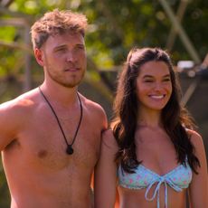 a shirtless jimmy p stands neck to a smiling ally in a bikini in a still from perfect match season 4