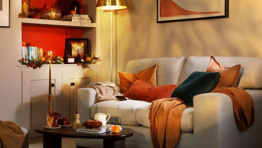 A neutral sofa is layered with warm autumnal cushions and throws, while soft golden lighting and pumpkin-filled shelves create a cosy, seasonal corner.